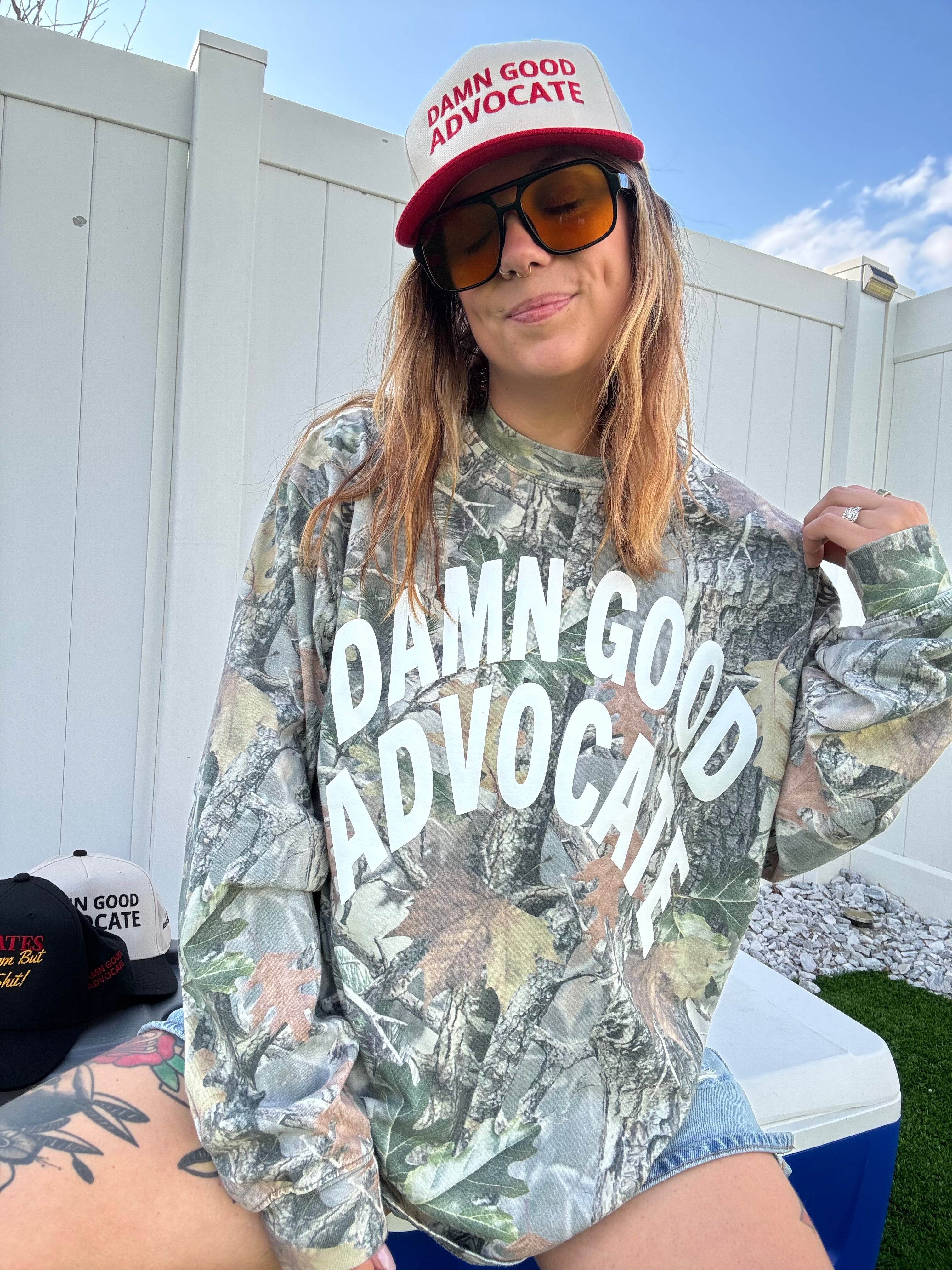 Person wearing a camouflage shirt and cap with 'Damn Good Advocate' text, standing outdoors.